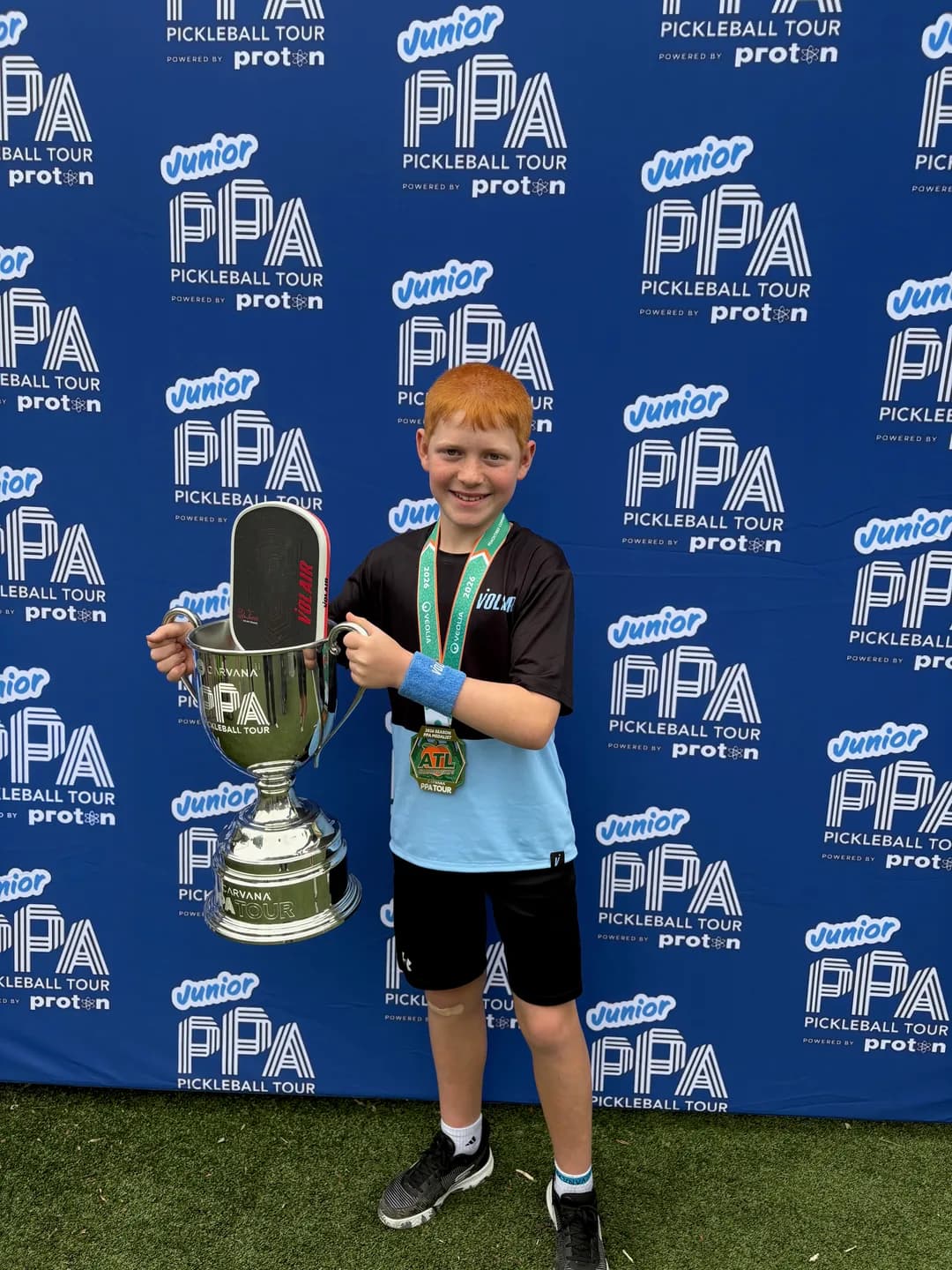 A young player holding a paddle and trophy beside a tournament backdrop