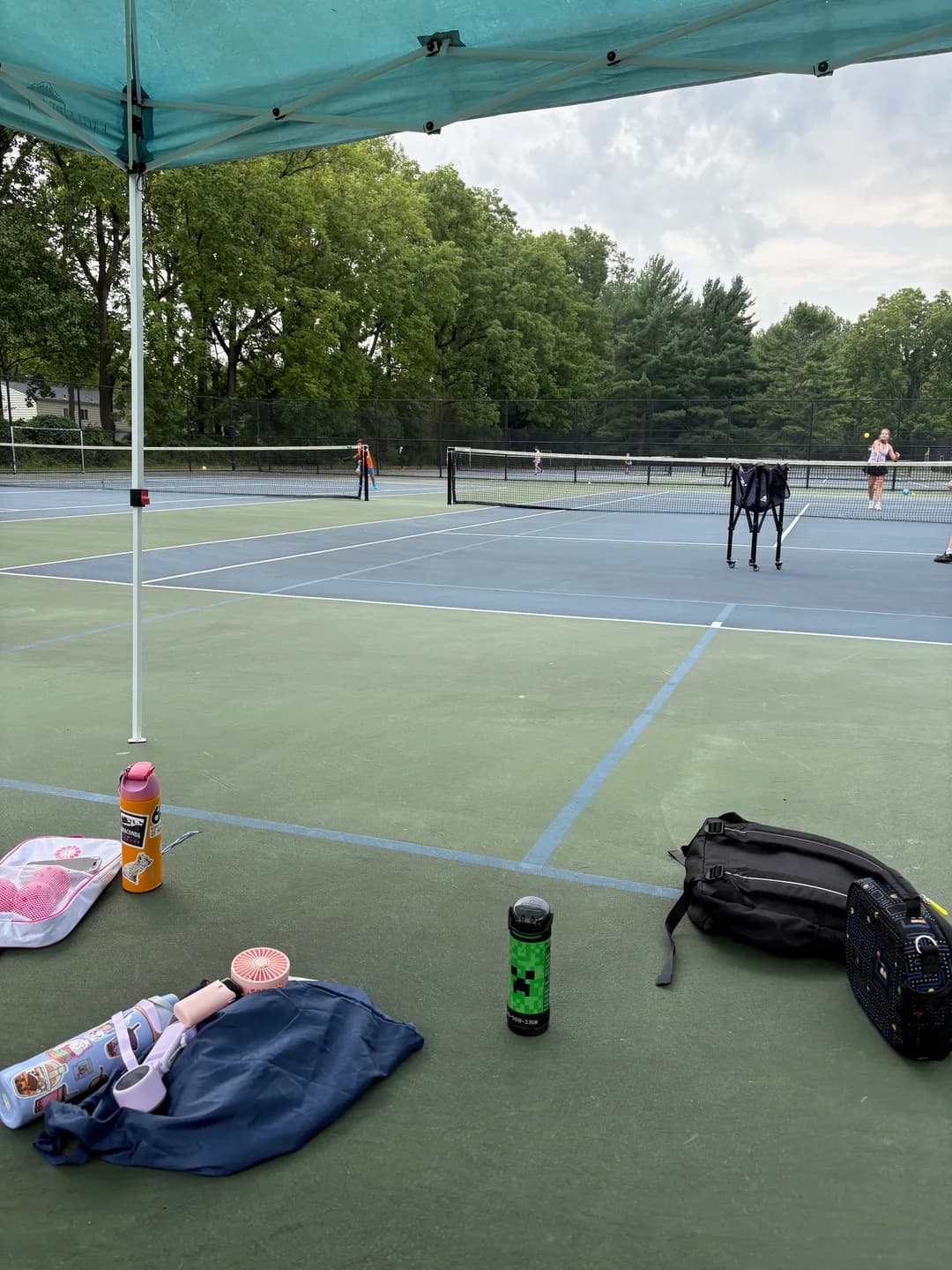 Bags and water bottles beside an outdoor court with players beyond the net