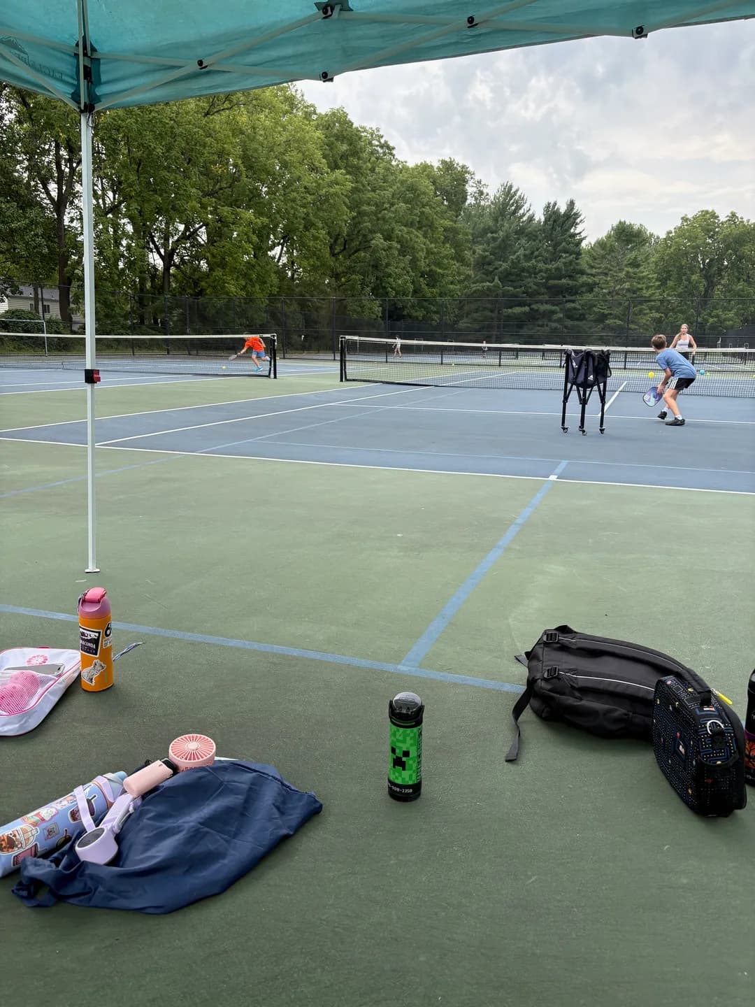 Bags and water bottles beside an outdoor court with players beyond the net
