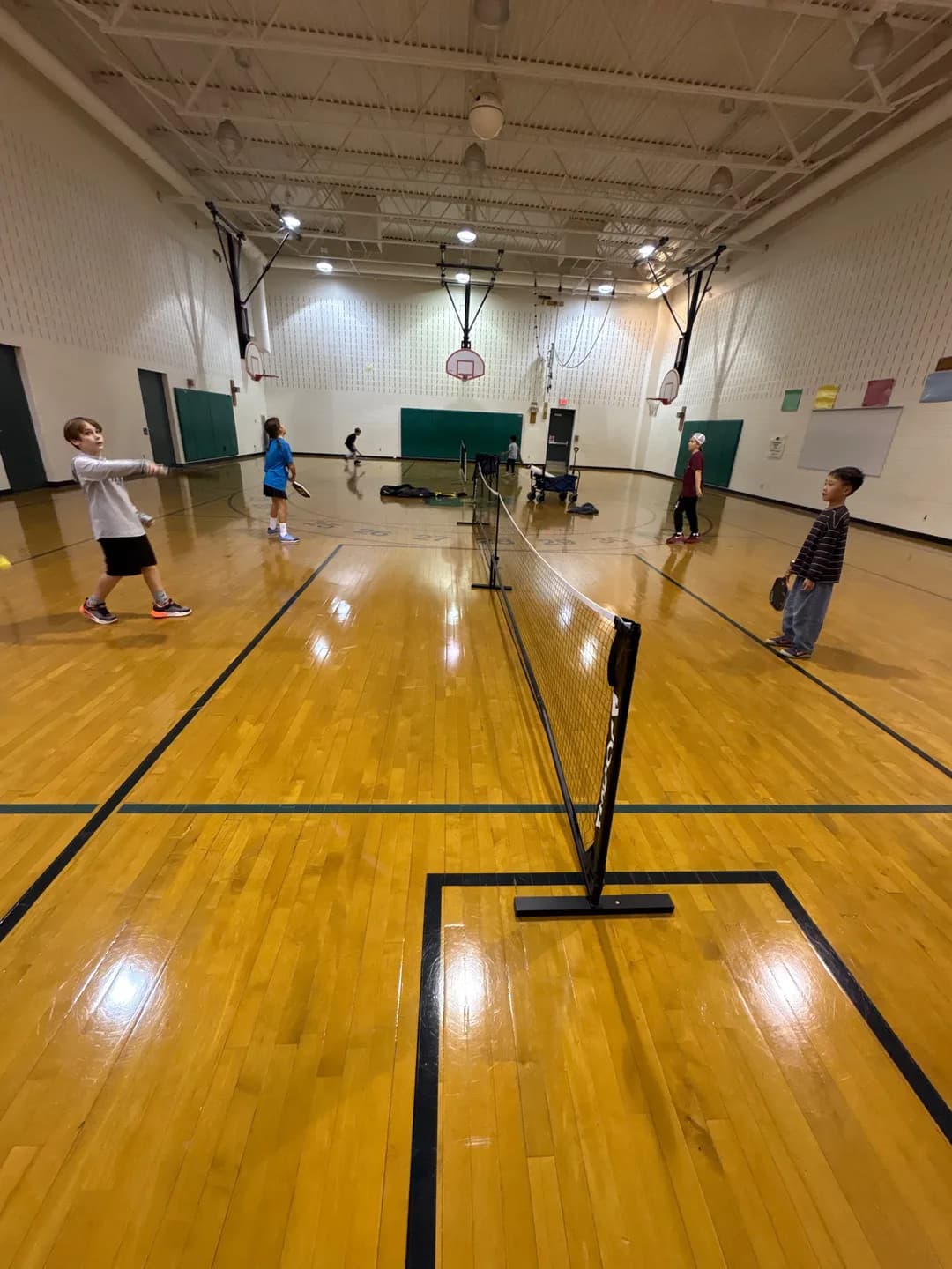 Young players on both sides of a pickleball net inside a gym