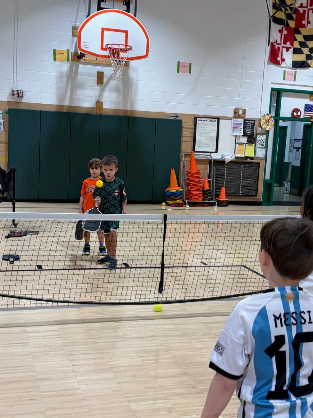 Young players with paddles playing pickleball across a portable net in a school gym
