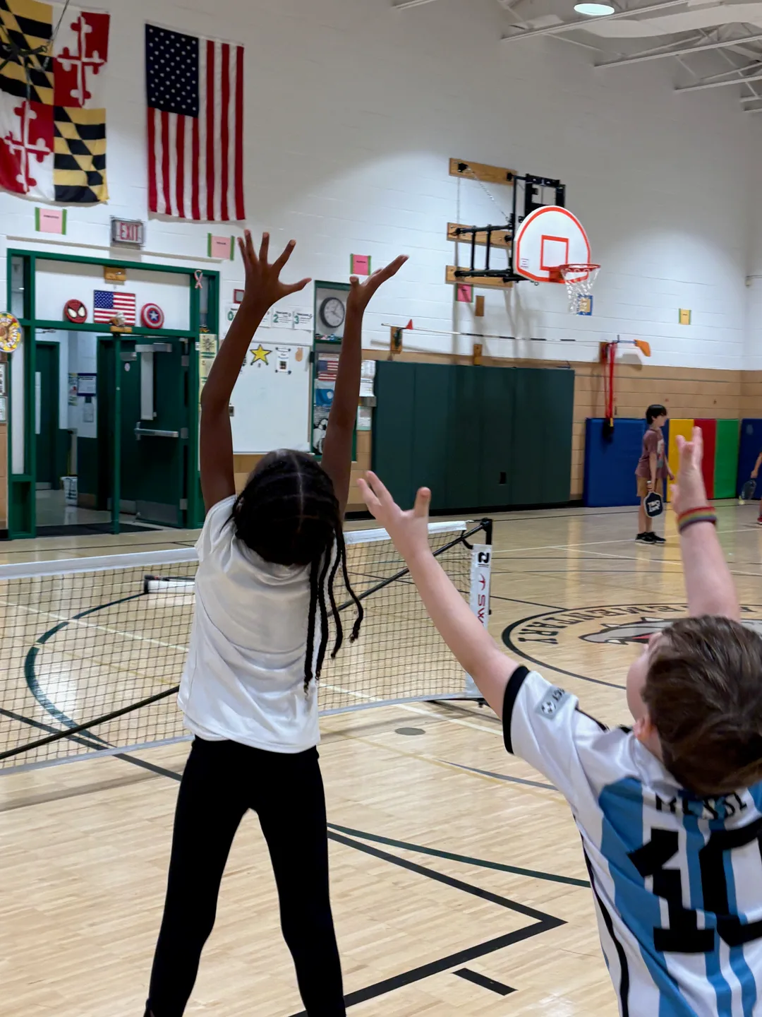 Young players raising their hands beside a pickleball net in a school gym